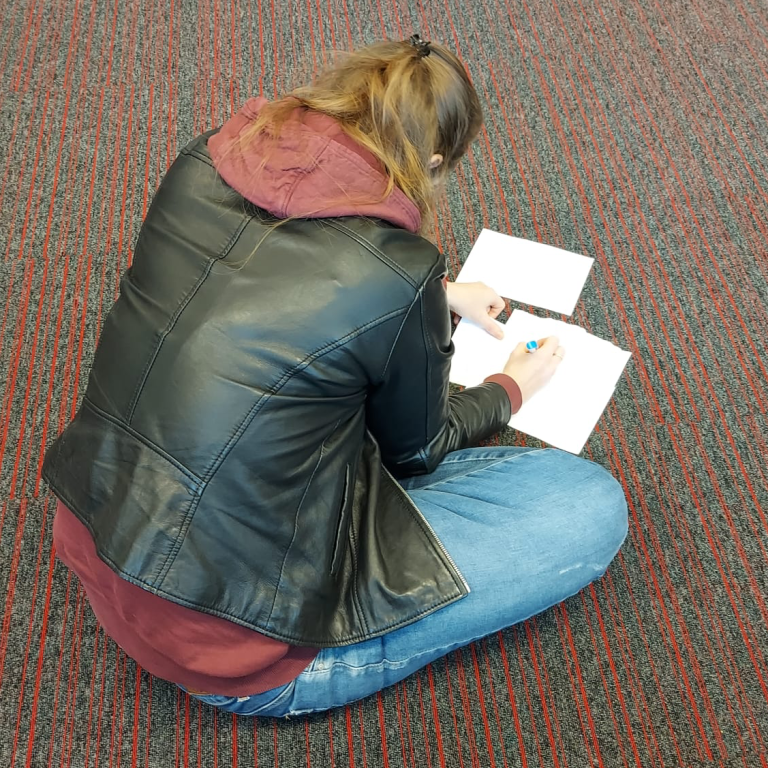 Christa zit op de grond en schrijft op witte blaadjes.